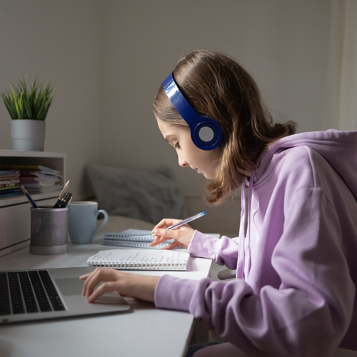 young girl in purple jumper and blue wireless headphones studying at laptop